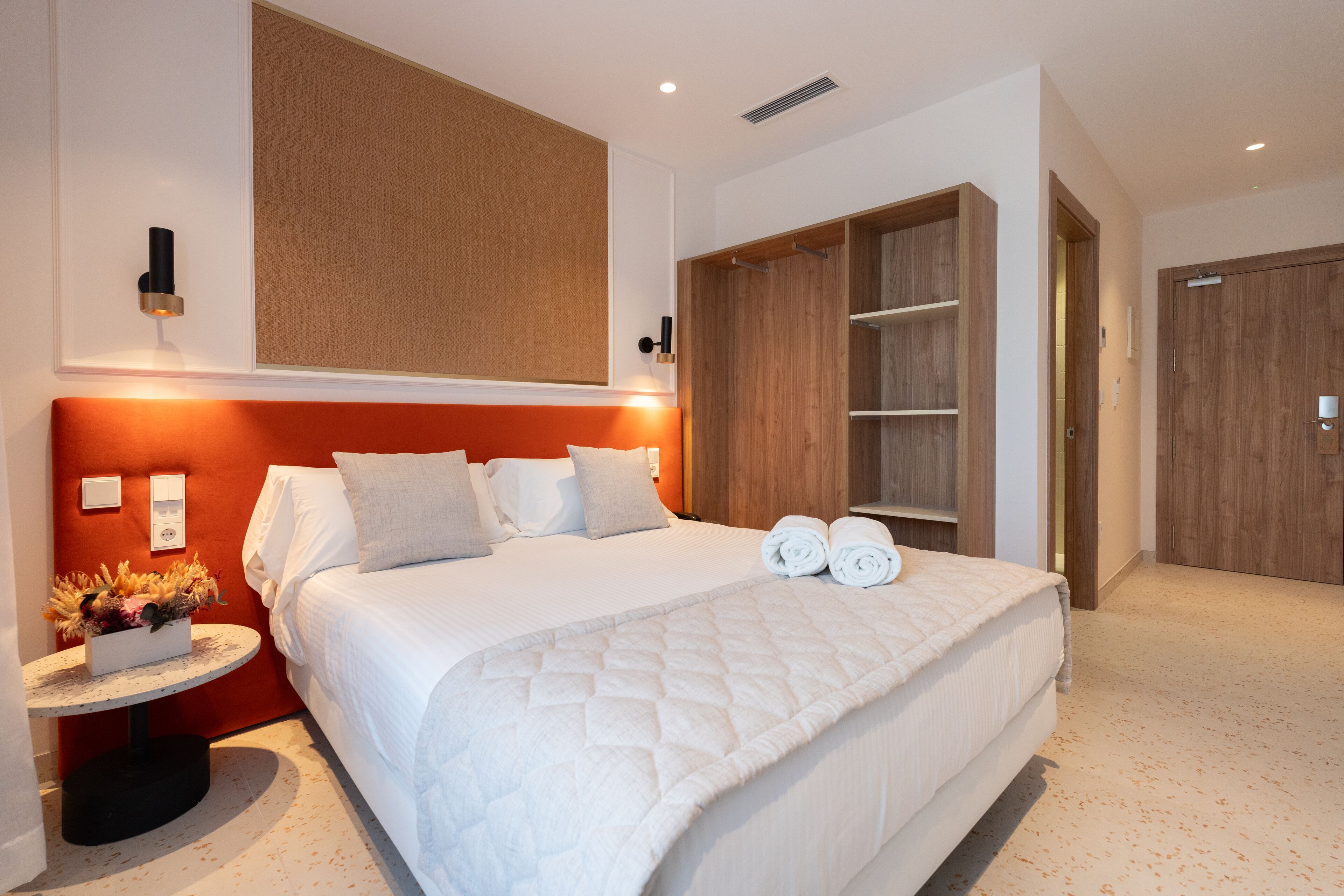 A modern hotel room with a large bed, a wooden wardrobe, and a small table with a flower arrangement.