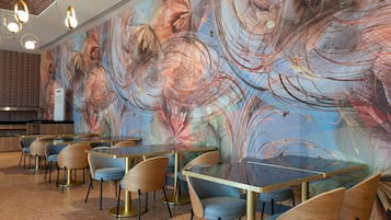 Interior de un restaurante moderno con mesas y sillas redondas, un mural abstracto grande en la pared y un piso moteado.