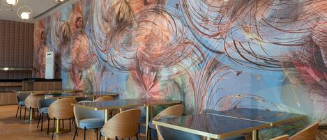 A modern restaurant interior with round tables and chairs, a large abstract mural on the wall, and a speckled floor.