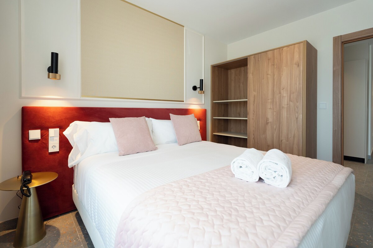 A hotel room with a large bed, a wooden wardrobe, a bedside table, and a wall-mounted light fixture.