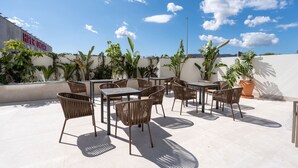 Un pati exterior amb taules i cadires, envoltat de plantes en testos i un cel blau clar.