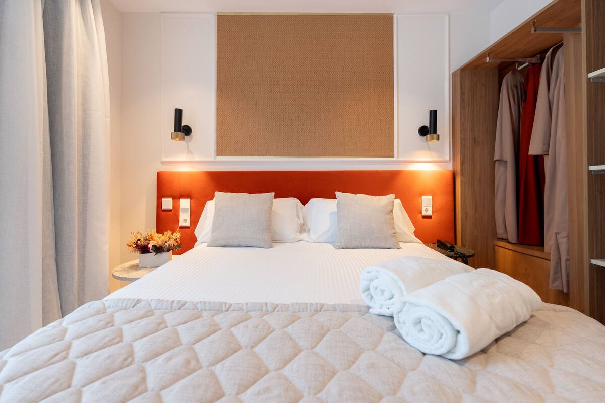 A neatly made bed with a quilted cover, a wall-mounted light fixture, and a wooden wardrobe with hanging clothes.