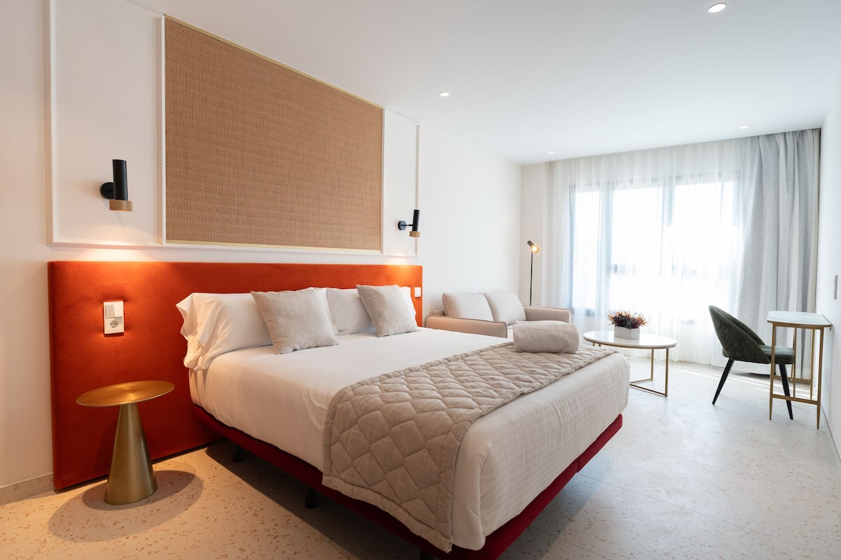 A modern hotel room with a large bed, a small round table, a chair, and a view of the city through large windows.