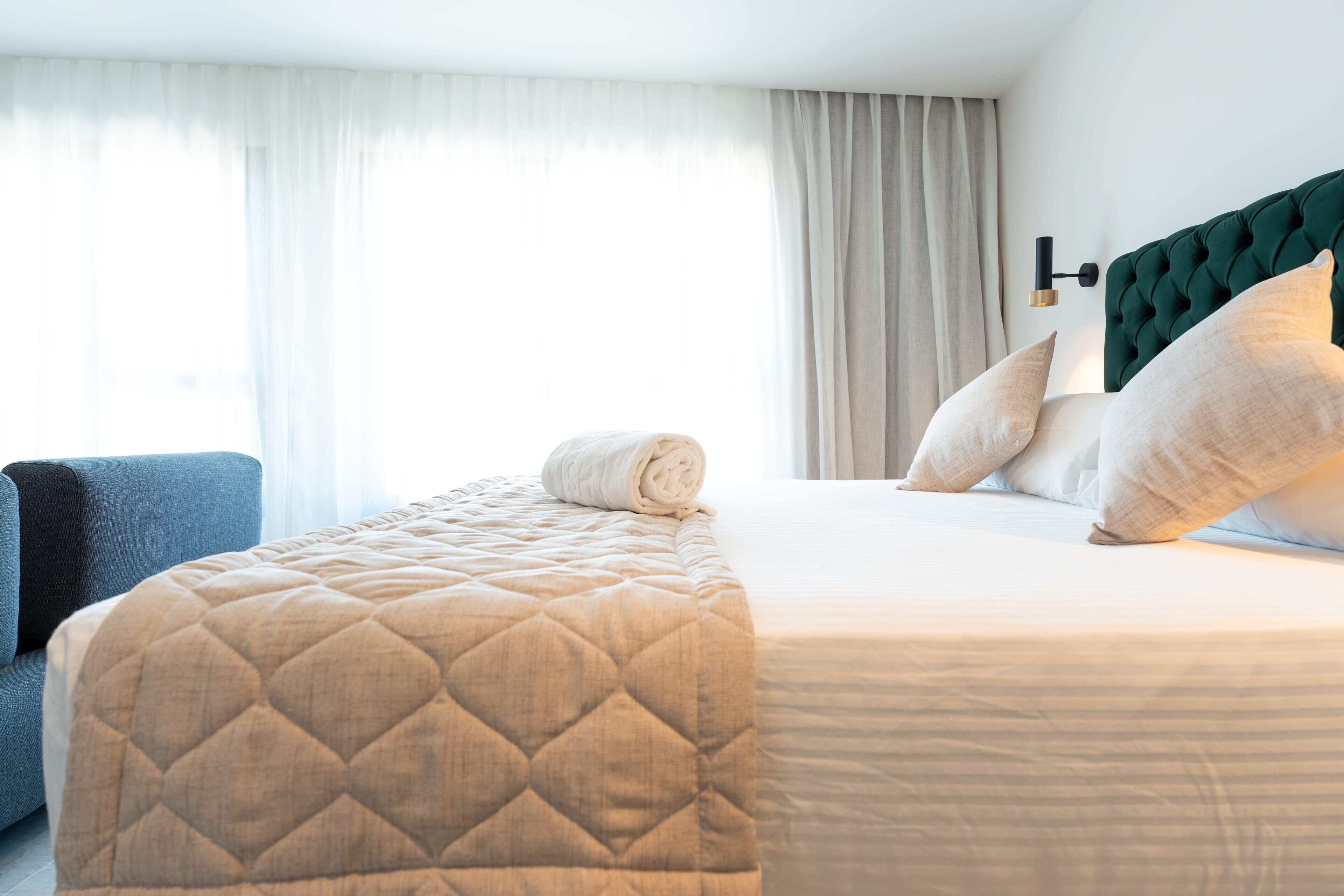 A hotel room with a bed, a blue armchair, and a window with curtains.