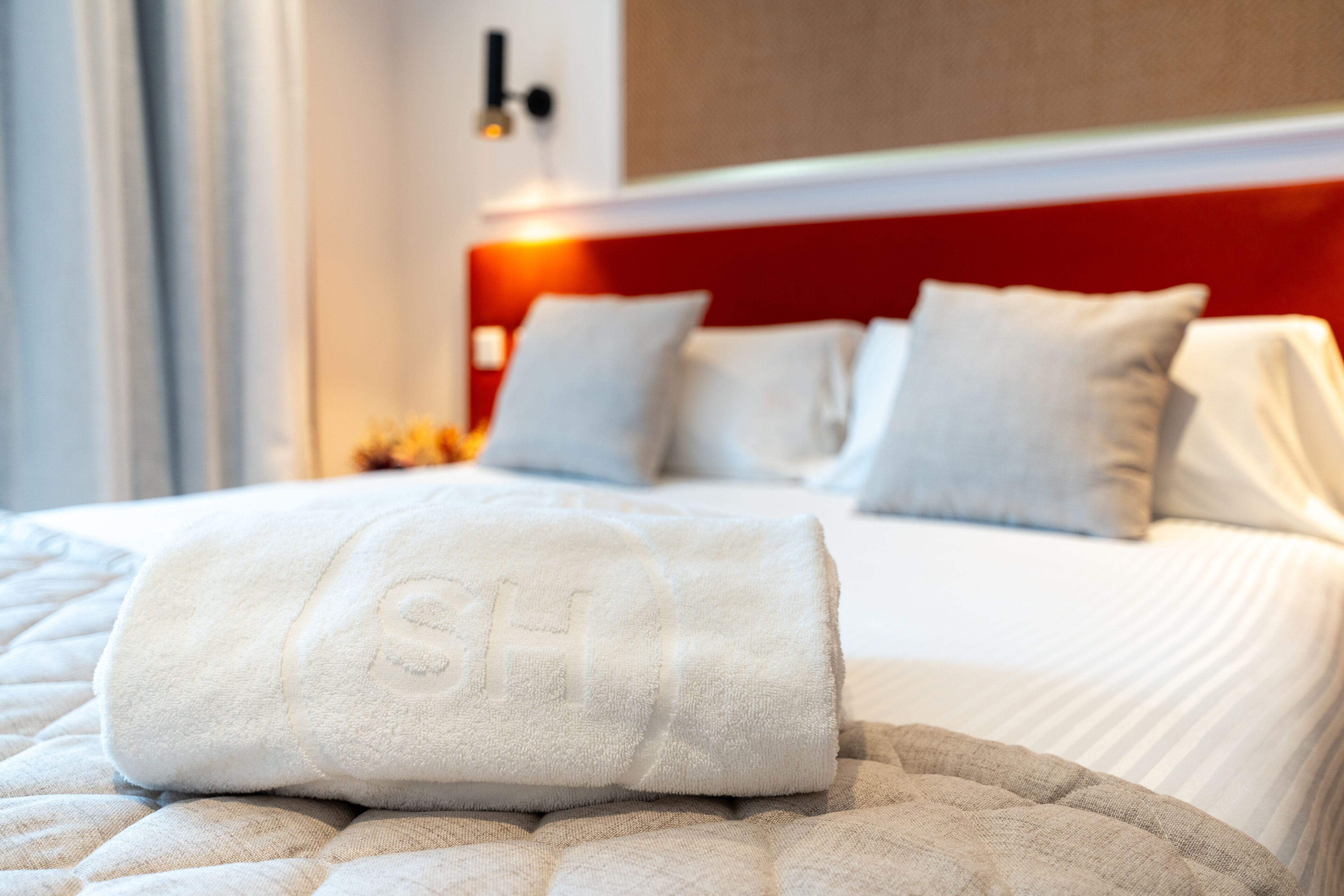 Ein ordentlich gemachtes Bett mit weißer Bettwäsche und Kissen, ein aufgerolltes weißes Handtuch auf dem Bett und ein rotes Kopfteil.