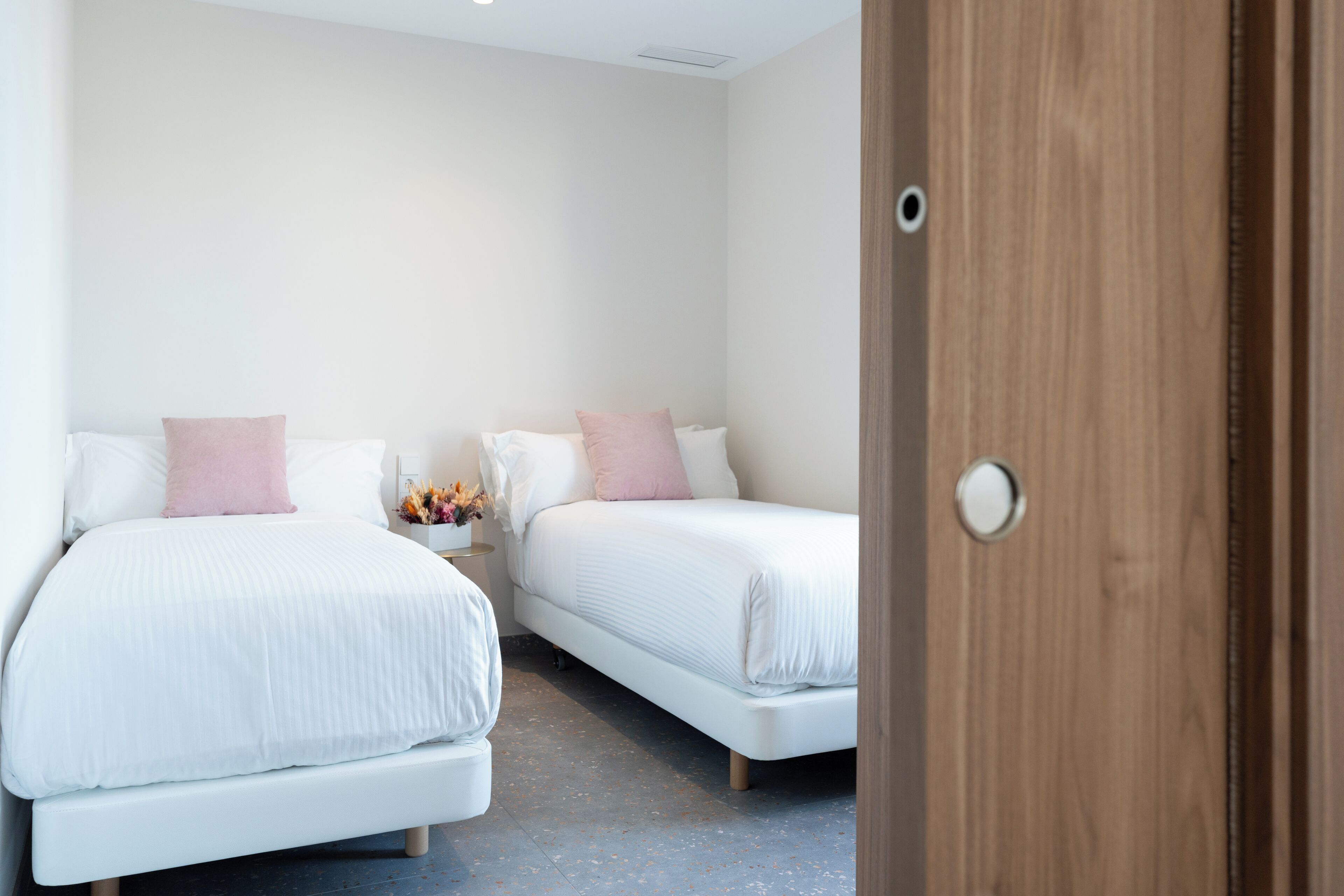 Une chambre d’hôtel avec deux lits, chacun étant doté de draps blancs et d’oreillers roses. On aperçoit une porte en bois sur le côté droit de l’image.