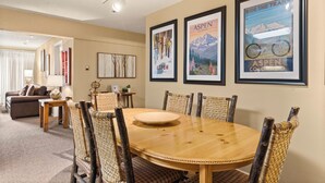 A dining area with a round wooden table and wicker chairs, framed posters on the wall, and a sofa in the background. - North Of Nell, Unit 2k (Aspen)