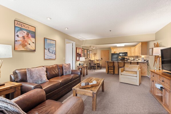 A spacious living room with a brown leather sofa, a wooden coffee table, and a beige armchair. A kitchen with wooden cabinets and a microwave is visible in the background.