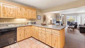 A kitchen with wooden cabinets, a dishwasher, and a large island. There is a living area with a couch and a fireplace. - North Of Nell, Unit 2k (Aspen)