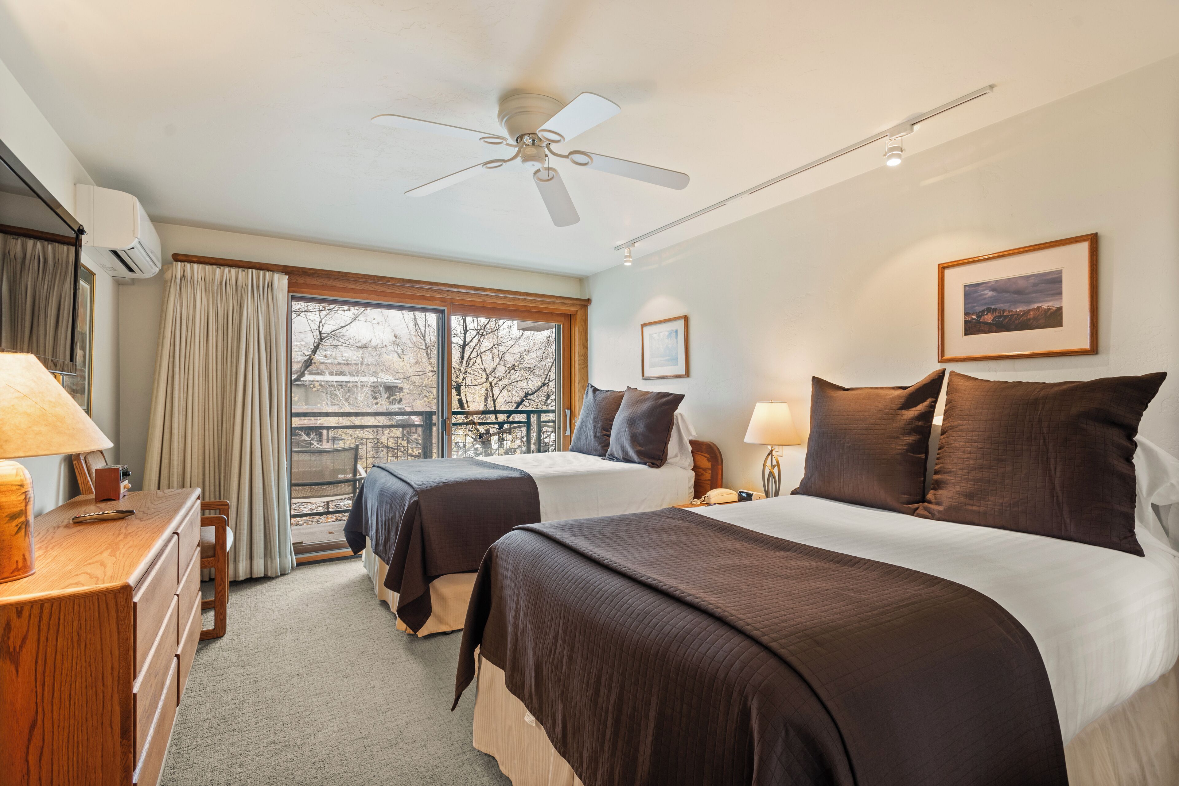 Une chambre d’hôtel avec deux lits, un ventilateur de plafond, une grande fenêtre avec des rideaux, une commode en bois et un tableau encadré au mur.