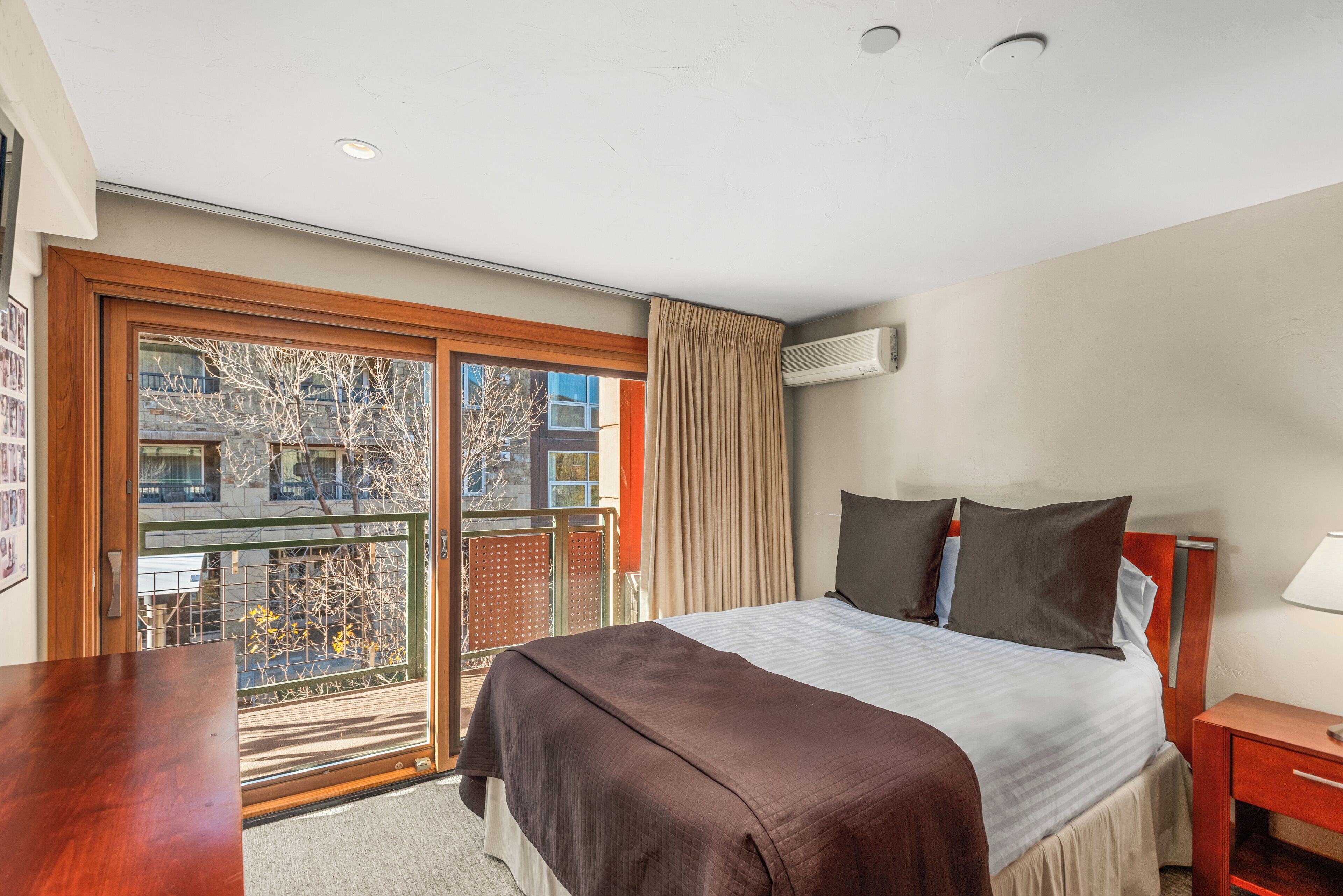 A hotel room with a large bed, a wooden desk, a window with a view of buildings and trees, and an air conditioning unit on the wall.