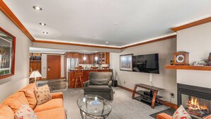 A modern living room with a sofa, coffee table, fireplace, TV, and kitchen in the background. - North Of Nell, Unit 3f (Aspen)