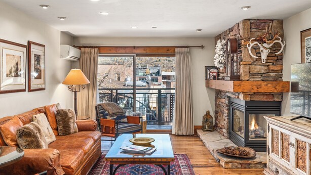 A cozy living room with a stone fireplace, leather sofa, and a view of the city through large windows. - North Of Nell, Unit 3i (Aspen)