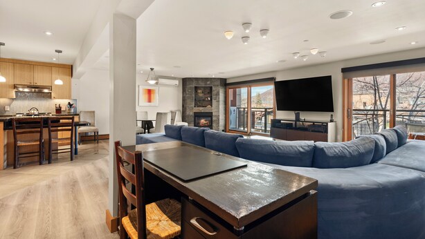 A modern kitchen with wooden cabinets, a dining area with a large table and chairs, a blue sofa, and a fireplace. - North Of Nell, Unit 3j (Aspen)