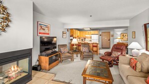 A living room with a fireplace, a television, and a kitchen in the background. - North Of Nell, Unit 3L (Aspen)