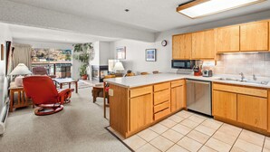 A modern kitchen with wooden cabinets, a dishwasher, and a large island. There is a dining area with a red chair and a table, and a view of a balcony with plants. - North Of Nell, Unit 3L (Aspen)