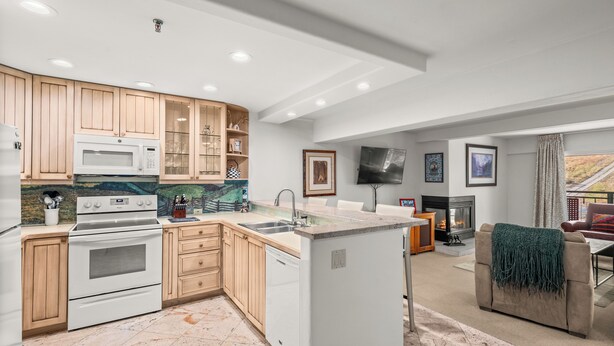 A modern kitchen with wooden cabinets, a built-in microwave, and a dishwasher. There is a seating area with a sofa and a fireplace.