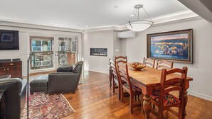 A dining area with a wooden table, chairs, and a chandelier. There is a large window with a view of trees and a fireplace in the background. - North Of Nell, Unit 4e (Aspen)