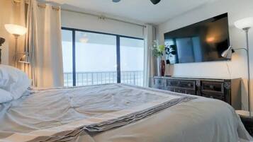 A hotel room with a large bed, a ceiling fan, a flat-screen TV, and a view of the sea through a large window.
