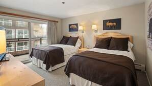 A hotel room with two beds, a desk, and a balcony with a view of buildings.