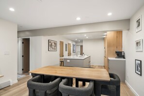 Eine moderne Küche mit einer zentralen Kochinsel, einem Essbereich mit Holztisch und grau gepolsterten Stühlen sowie einem Flur zu einer Tür.