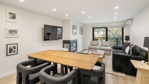 A modern living room with a wooden dining table, grey upholstered chairs, a black sofa, a fireplace, and a large TV mounted on the wall. - North Of Nell, Unit 4h (Aspen)