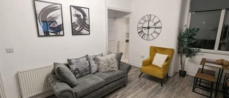 Een woonkamer met een grijze bank, een gele fauteuil en een ronde wandklok.