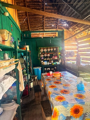 A rustic kitchen with wooden shelves, a table with a floral tablecloth, and a thatched roof. - Hospedaje  Reina Arriera Amazonas  (Leticia)