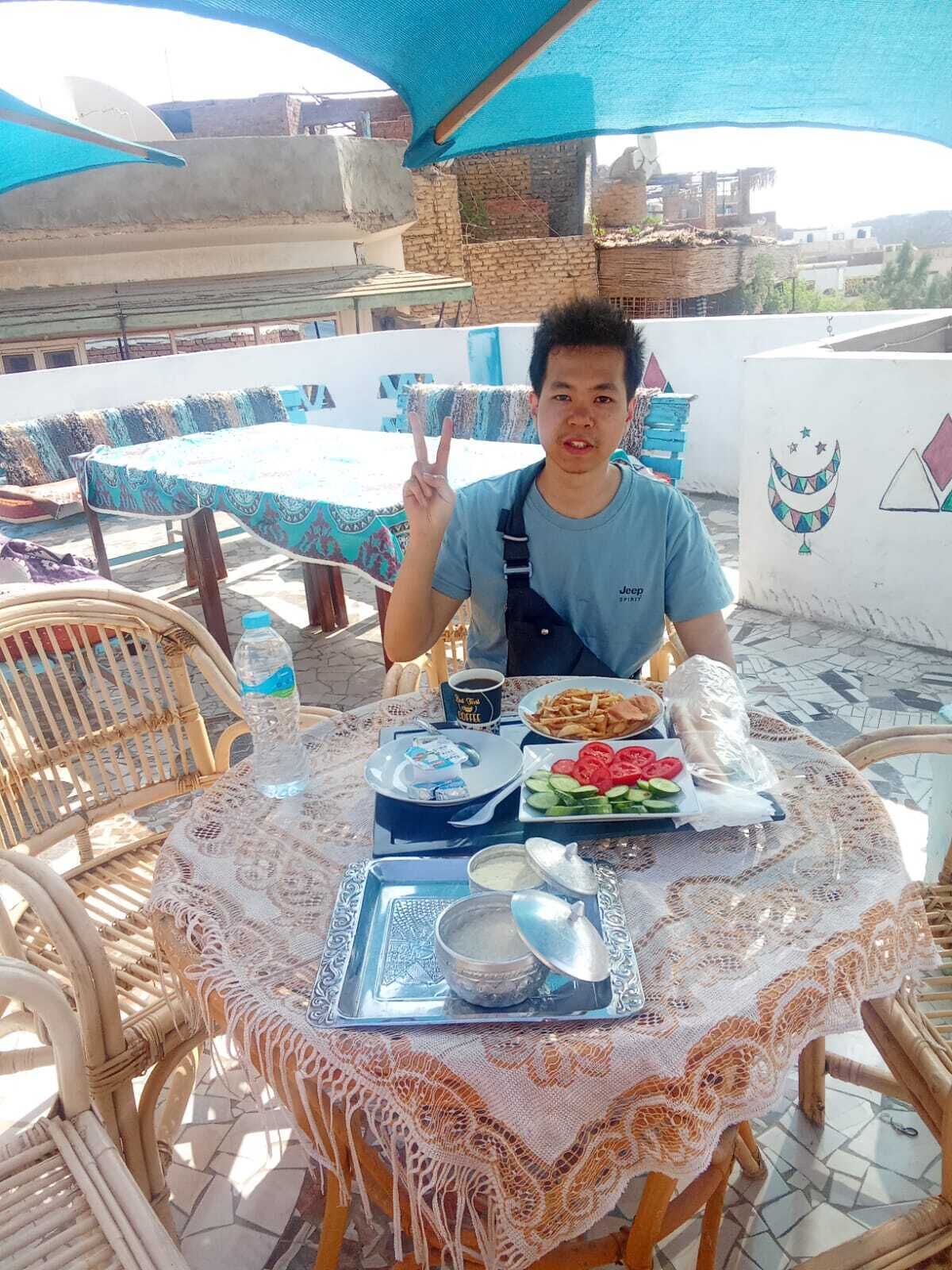 A person seated at a table with a variety of food items, including a plate of sliced tomatoes and cucumbers, a bowl of dip, and a plate of fries. The table is covered with a lace tablecloth, and there are wicker chairs around it.