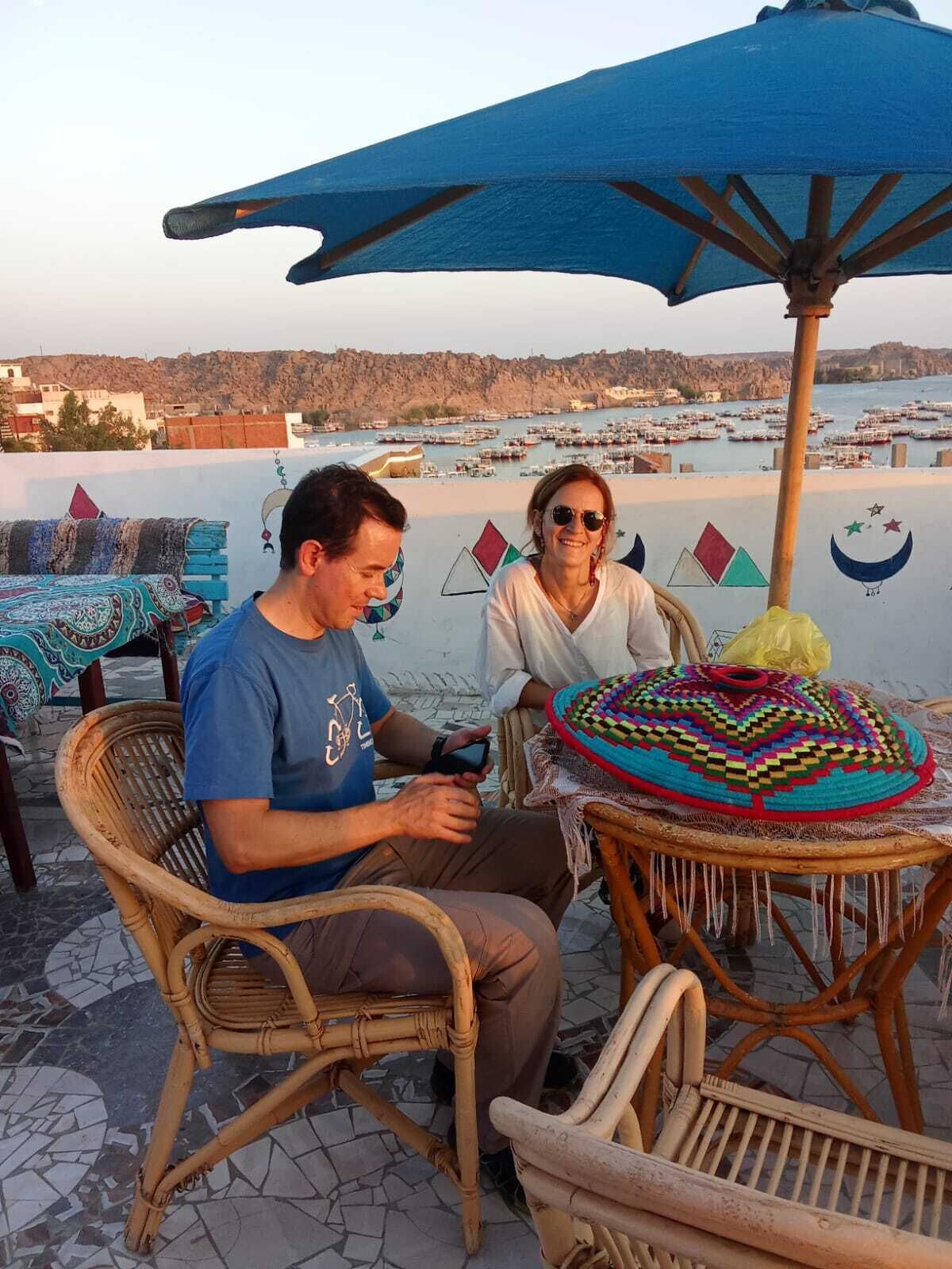 Two people seated at a table with a colorful woven basket, under a blue umbrella, overlooking a harbor with boats.