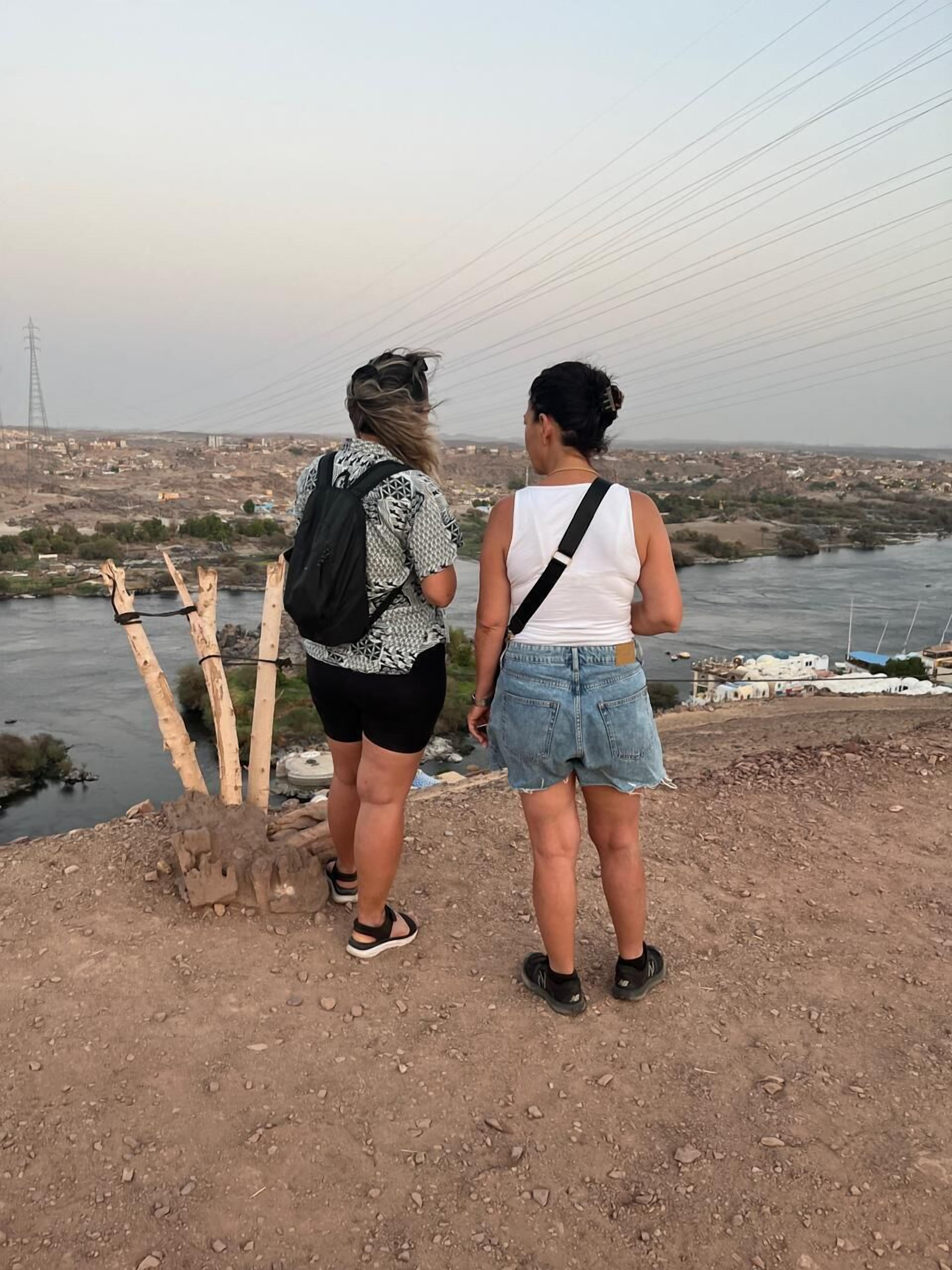 Two people standing on a rocky overlook with a river and distant buildings in the background.