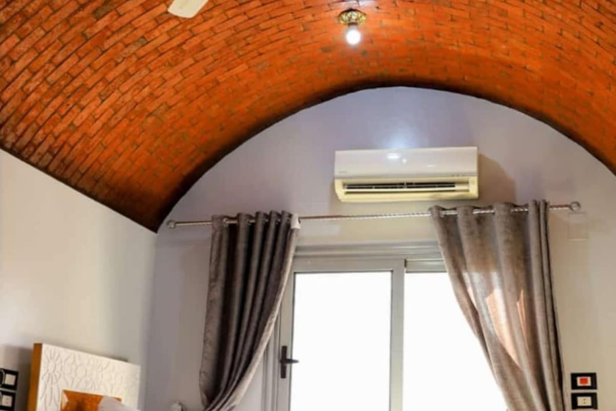 Una habitación de hotel con techo de ladrillo, unidad de aire acondicionado, cama con almohadas y ventana con cortinas.