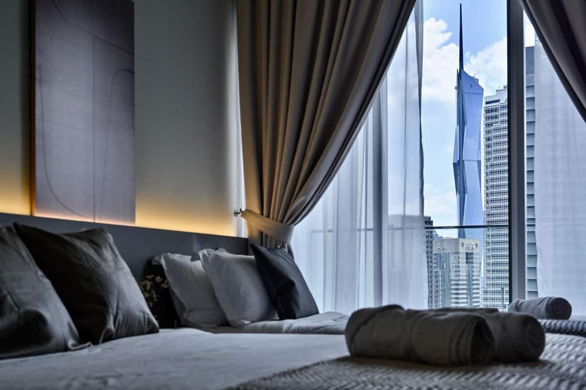 A modern hotel room with a large bed, a view of a city skyline, and a window with sheer curtains.