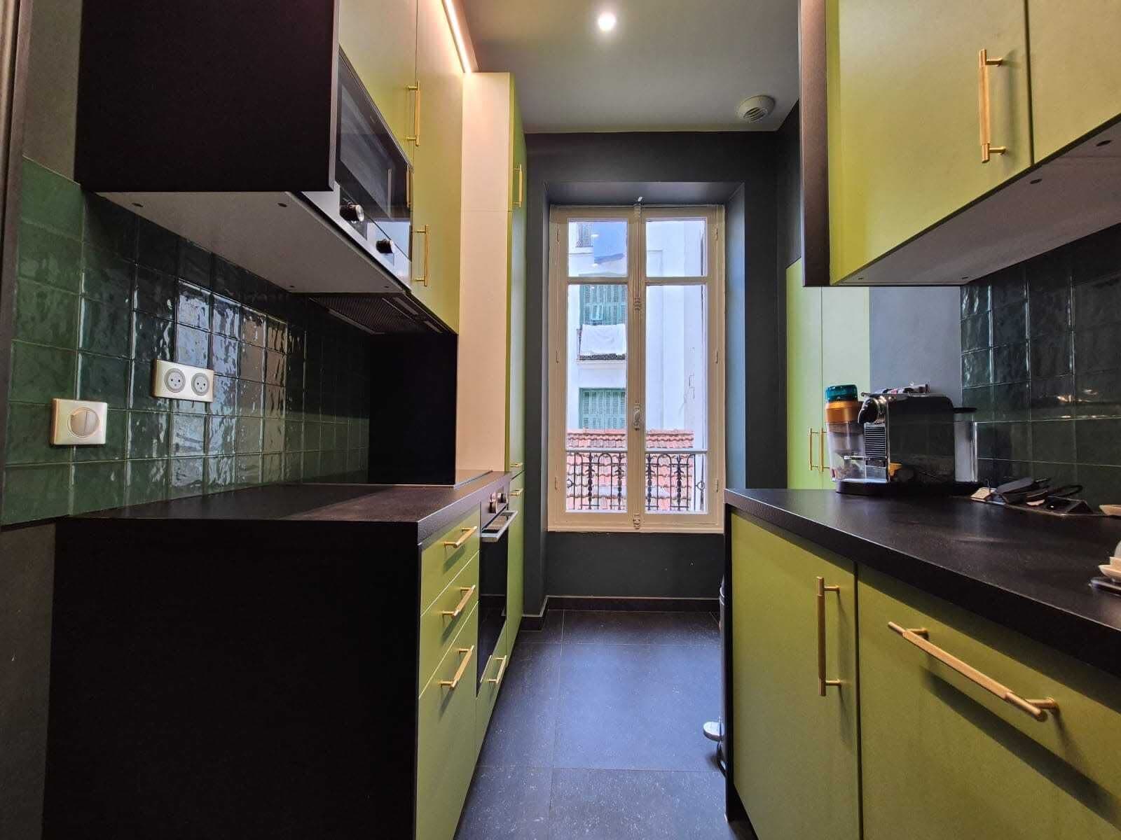 A narrow kitchen with green cabinets, a black countertop, and a window with a view of buildings.