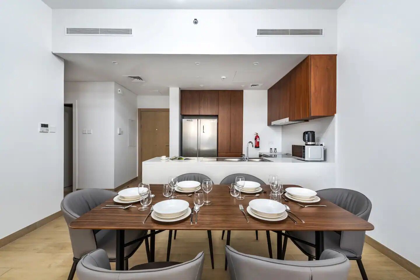 A modern dining area with a wooden table set for six, surrounded by grey chairs. A kitchen with wooden cabinets and stainless steel appliances is visible in the background.