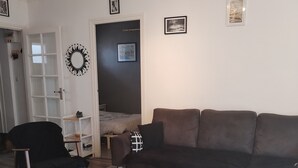 Un salon avec un canapé marron, un fauteuil noir, une table basse avec des magazines, et une porte ornée d’un miroir et de cadres de photos.