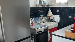 Une cuisine de taille réduite, équipée d’un réfrigérateur en acier inoxydable, d’une cuisinière et d’un évier. On peut y voir des aimants décoratifs sur le réfrigérateur, une horloge au mur et une fenêtre avec un rideau.