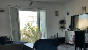 Un salon moderne comprenant un canapé, un fauteuil bleu, une table à manger avec des chaises, un téléviseur et un balcon offrant une vue sur des palmiers.
