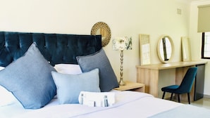 Een keurig opgemaakt bed met een gestoffeerd hoofdbord, een bureau met stoel, en een spiegel aan de muur.
