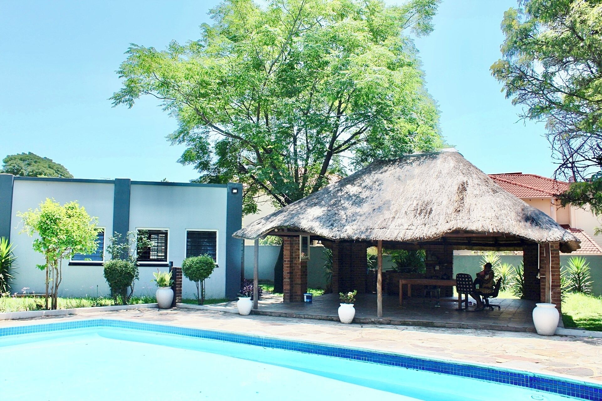 A pool area with a thatched-roof structure, a modern building, and surrounding greenery.