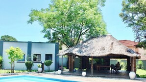 A pool area with a thatched-roof structure, a modern building, and surrounding greenery.