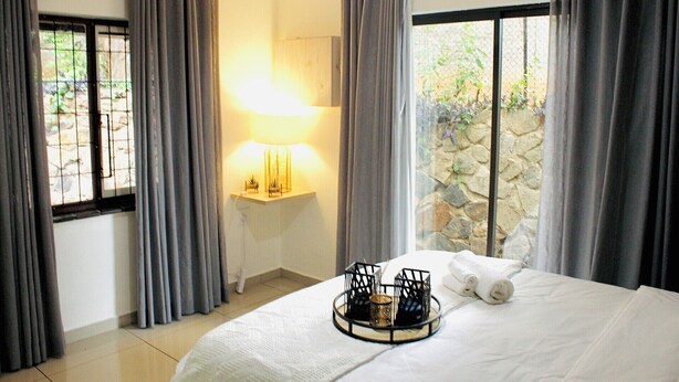A hotel room with a bed, a bedside lamp, a tray with towels, and a view of greenery through the window.