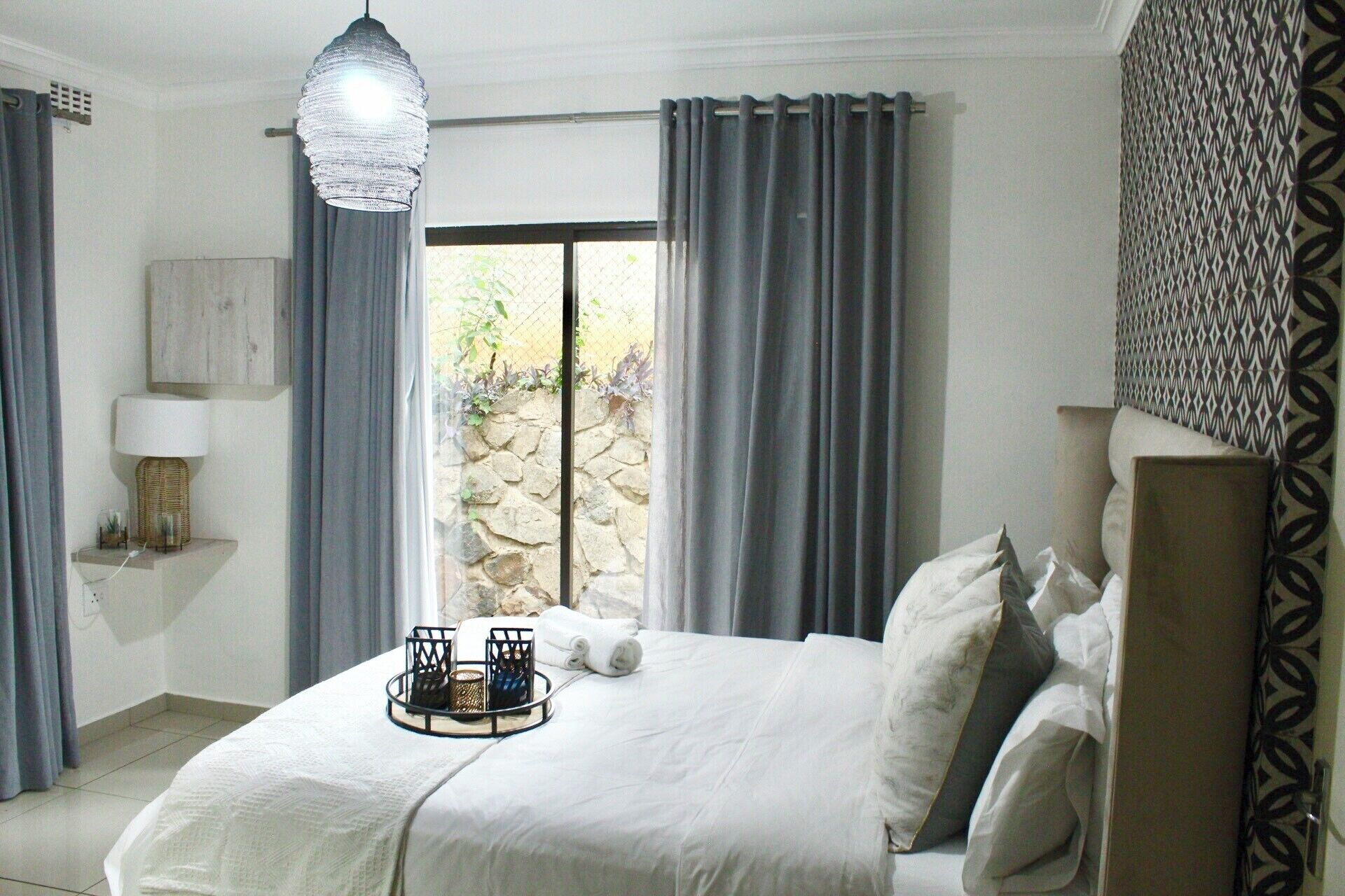 A bedroom with a bed, a bedside lamp, a decorative hanging light, and a view of the outdoors through a window.