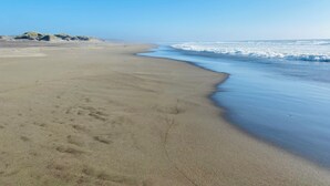 A sandy beach with gentle waves, a clear sky, and dunes in the distance.