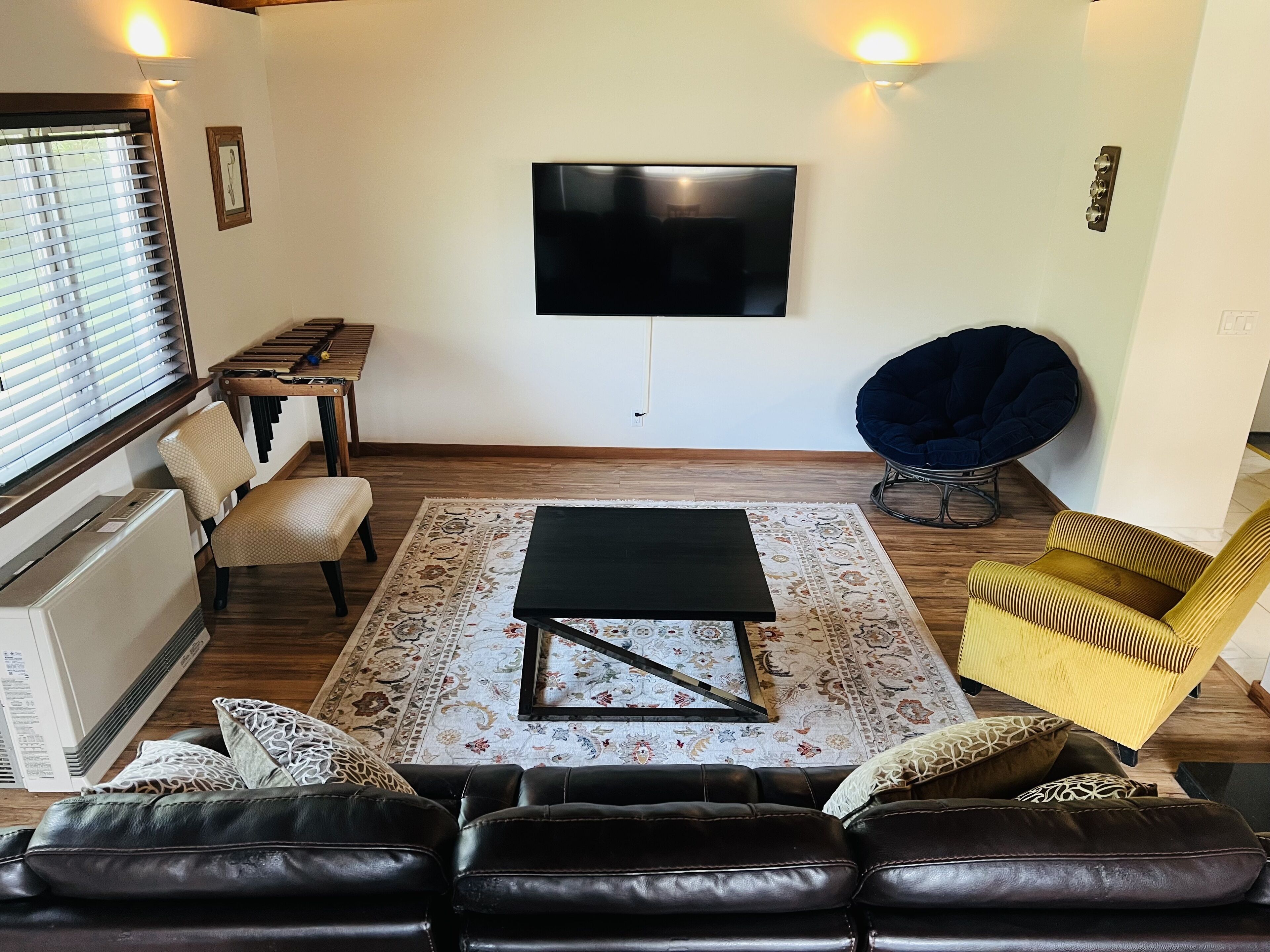 Un salon avec un canapé en cuir noir, une table basse, un tapis à motifs, un téléviseur à écran plat fixé au mur, une table à manger en bois avec des chaises, un fauteuil en forme de poire bleu et un fauteuil jaune.