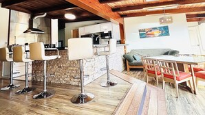 A spacious room with a high ceiling, wooden beams, a dining area with red chairs, and a kitchen area with white cabinets.