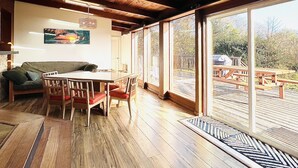 A spacious room with wooden floors, a dining table with chairs, a sofa, and large windows offering a view of an outdoor deck.
