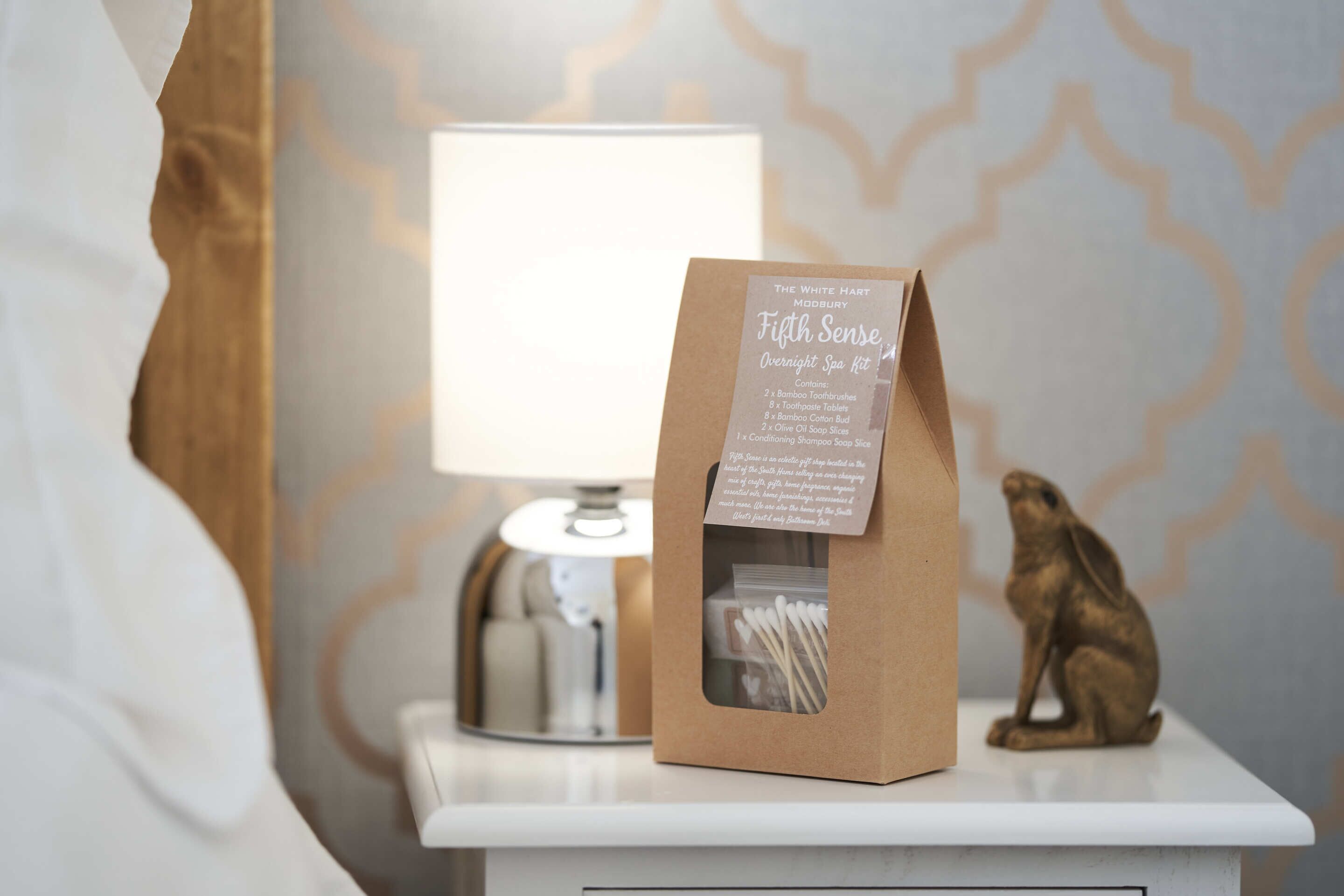 Una mesita de noche con una lámpara, una bolsa de papel marrón etiquetada “Fifth Sense Overnight Spa Kit” y una pequeña estatua dorada.