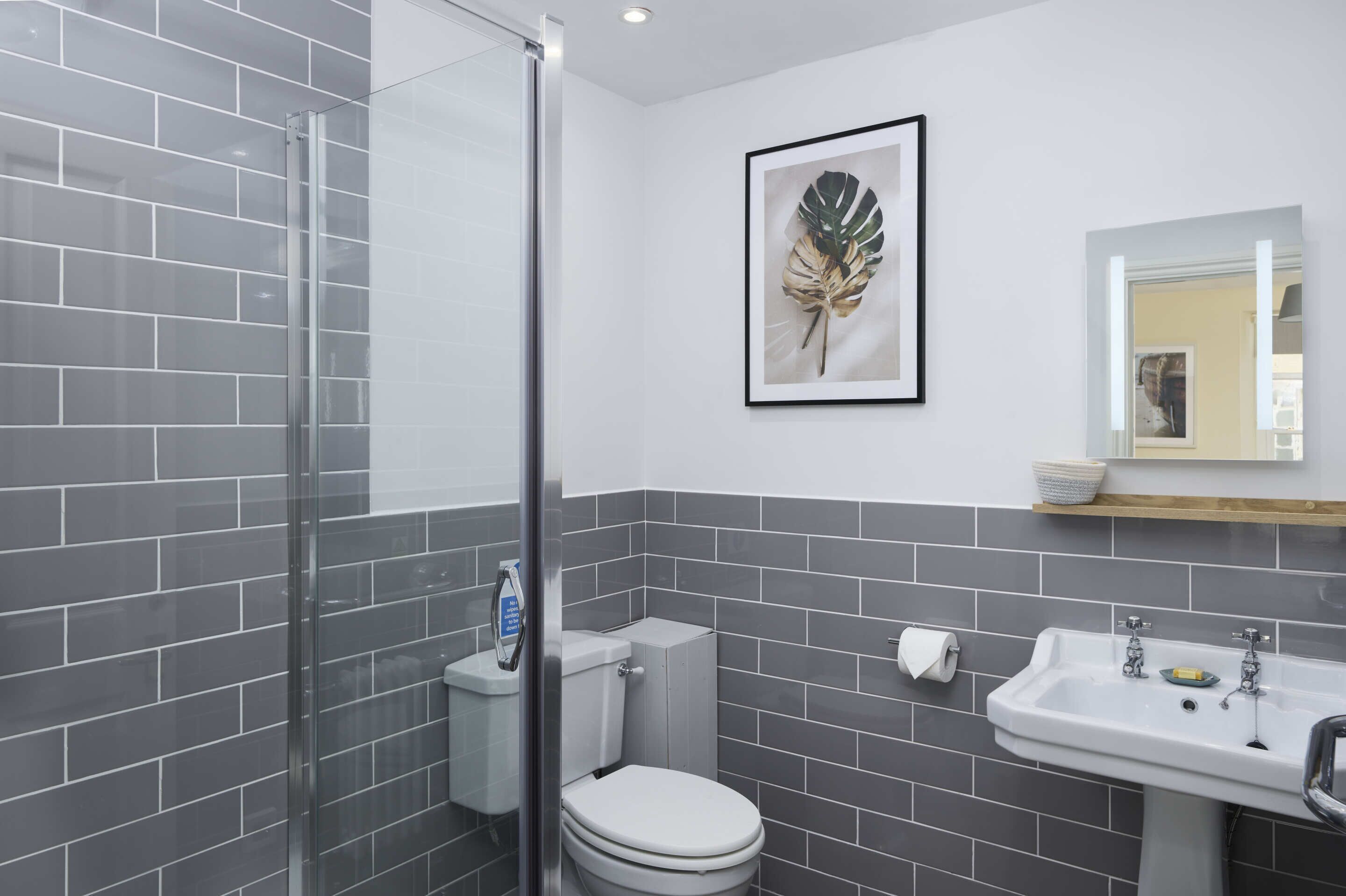 A bathroom with grey tiled walls, a glass shower enclosure, a white toilet, a sink with a mirror above it, and a framed leaf artwork on the wall.
