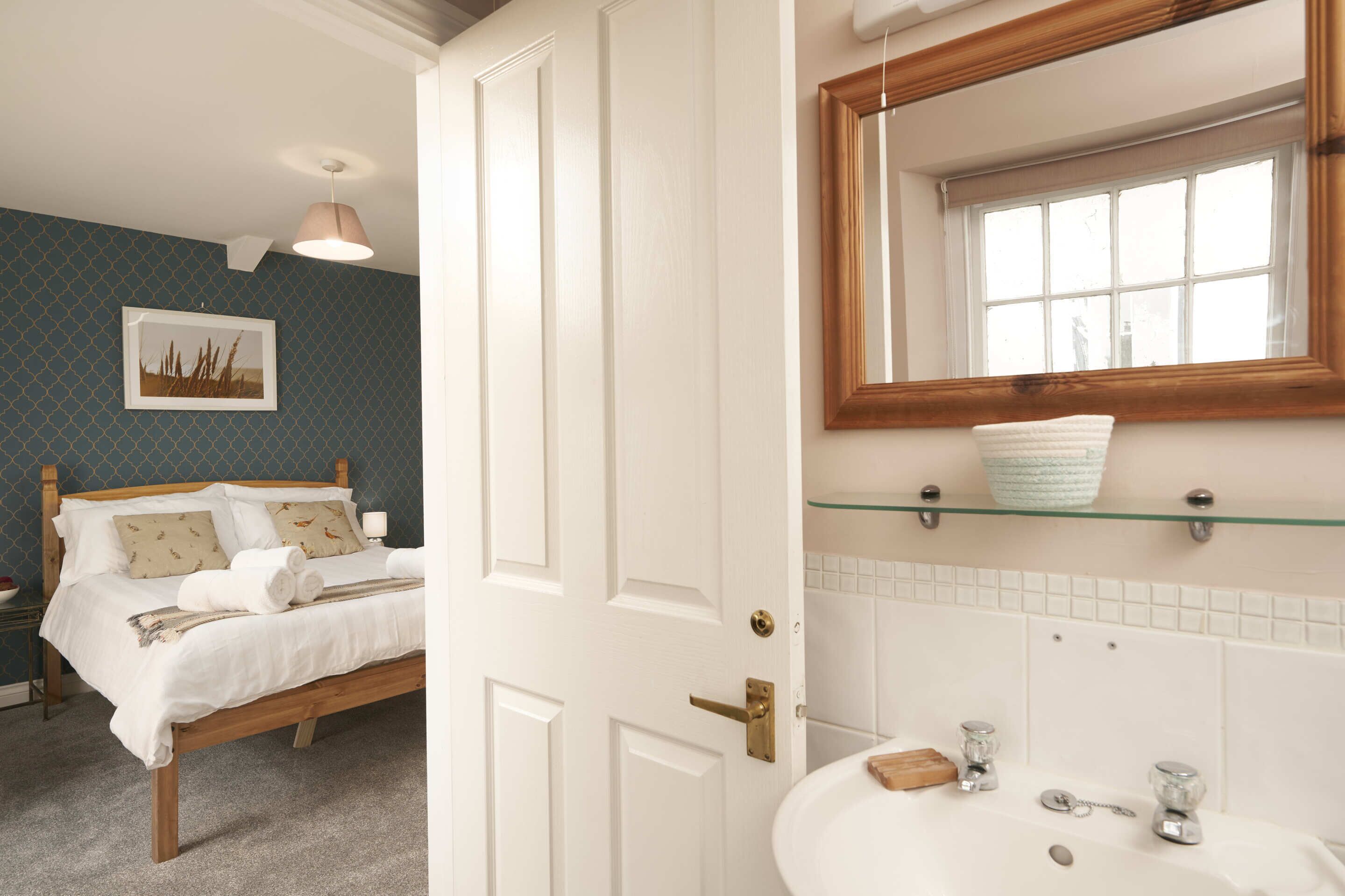 Une chambre à coucher avec un lit en bois, une table de chevet et une commode. La salle de bain est équipée d’un lavabo, d’un miroir et d’étagères.